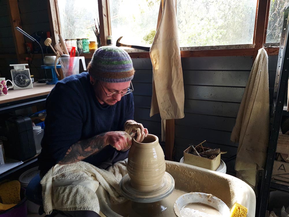 Stace Robertson sits at a pottery wheel in his studio, shaping a tall clay pot with his hands.